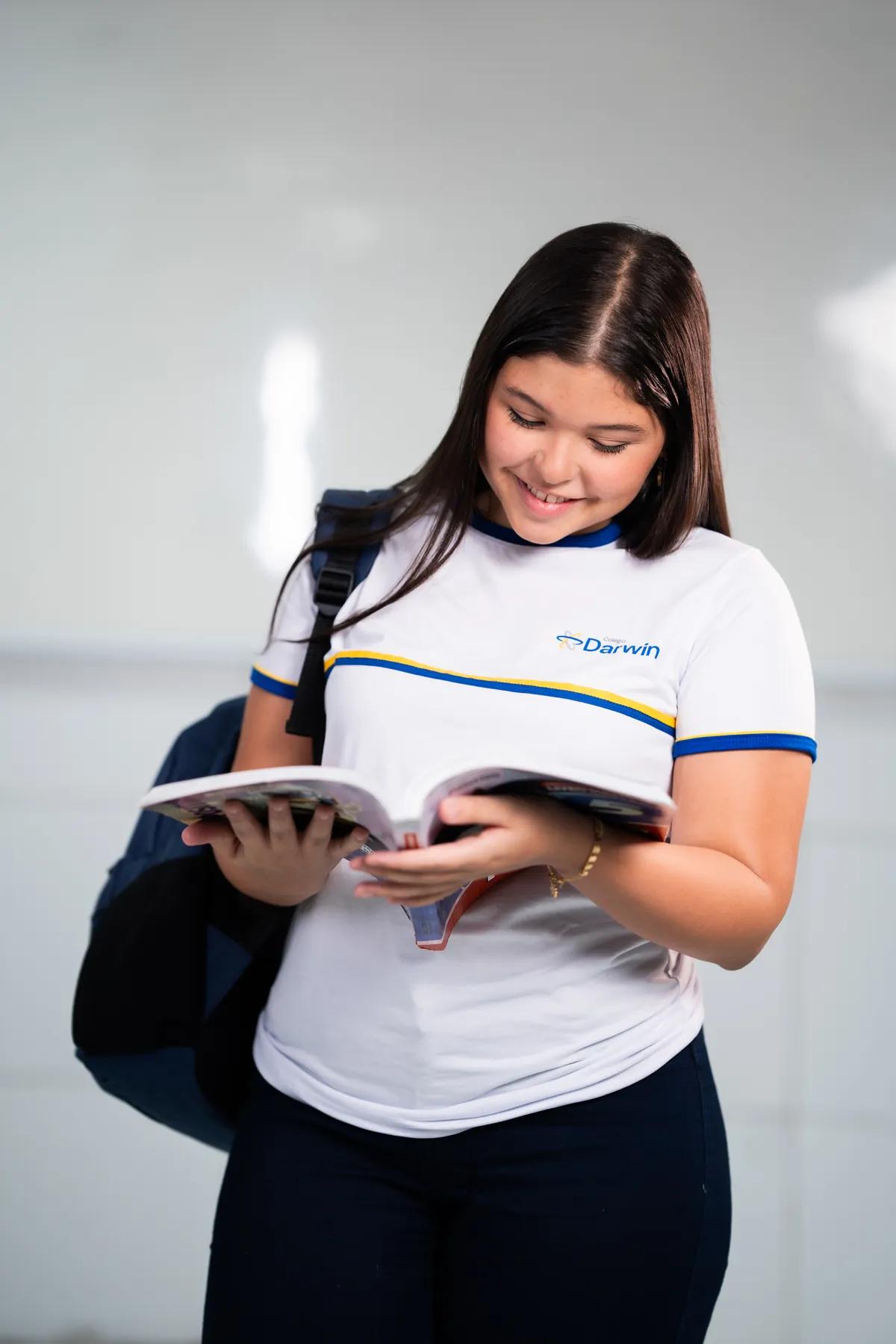 Aluna sorrindo enquanto lê livro didático em sala de aula.