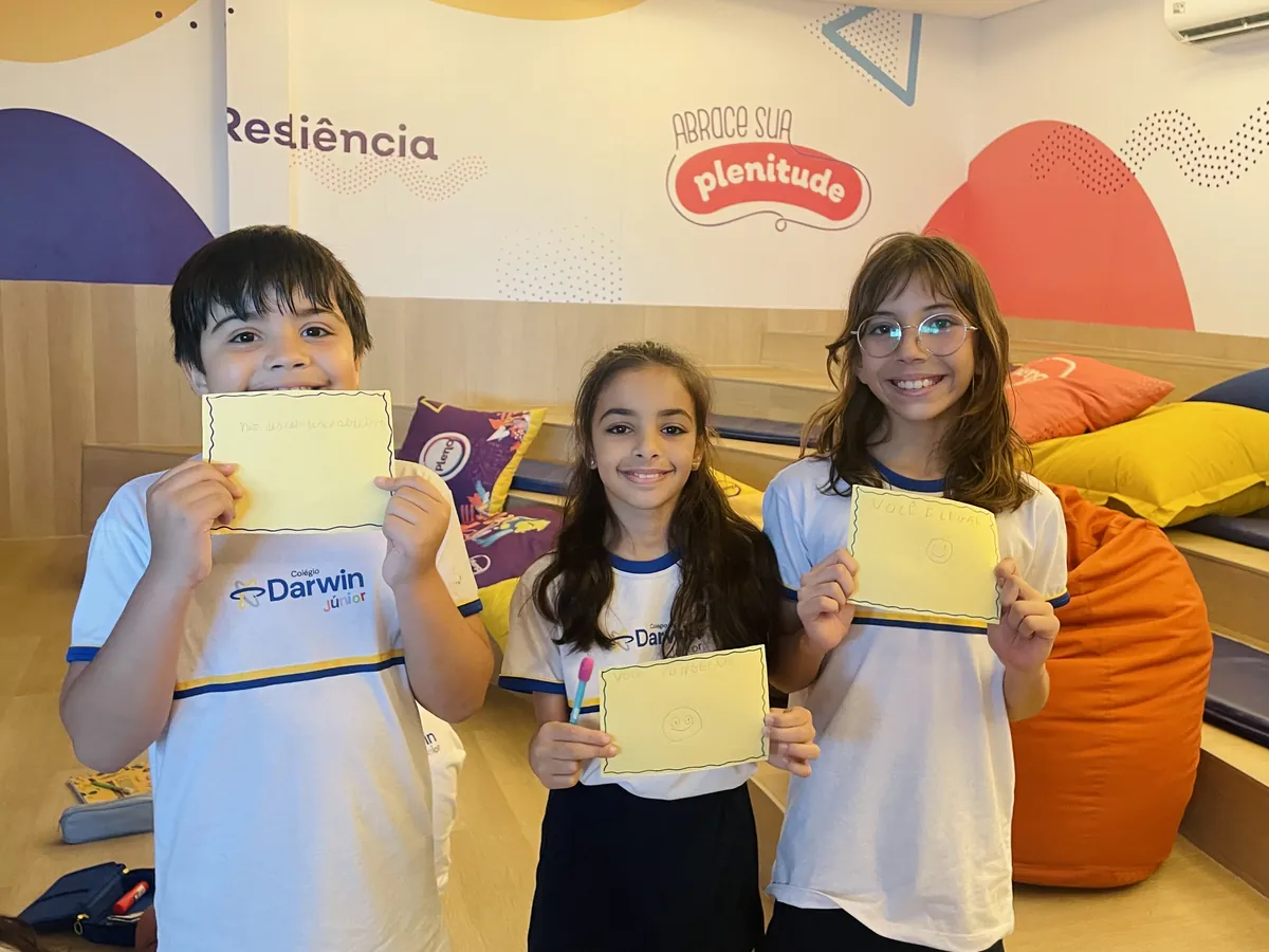 Alunos mostrando cartões amarelos em ambiente colorido de convivência.