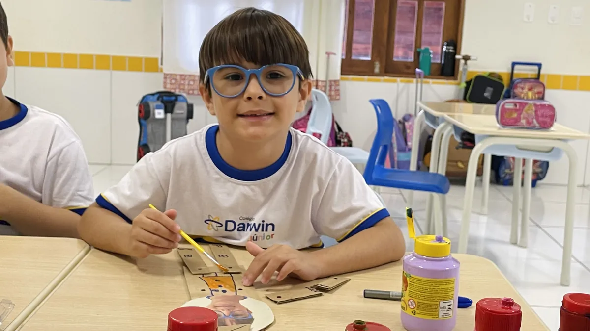 Menino de óculos azuis pintando figura de papelão em sala de aula.