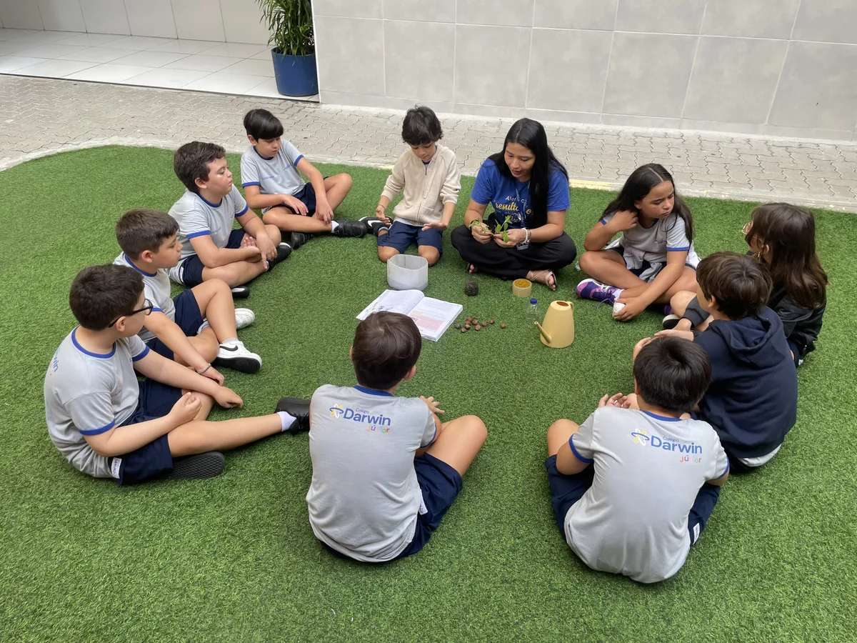 Turma sentada em roda com a professora em área externa gramada.