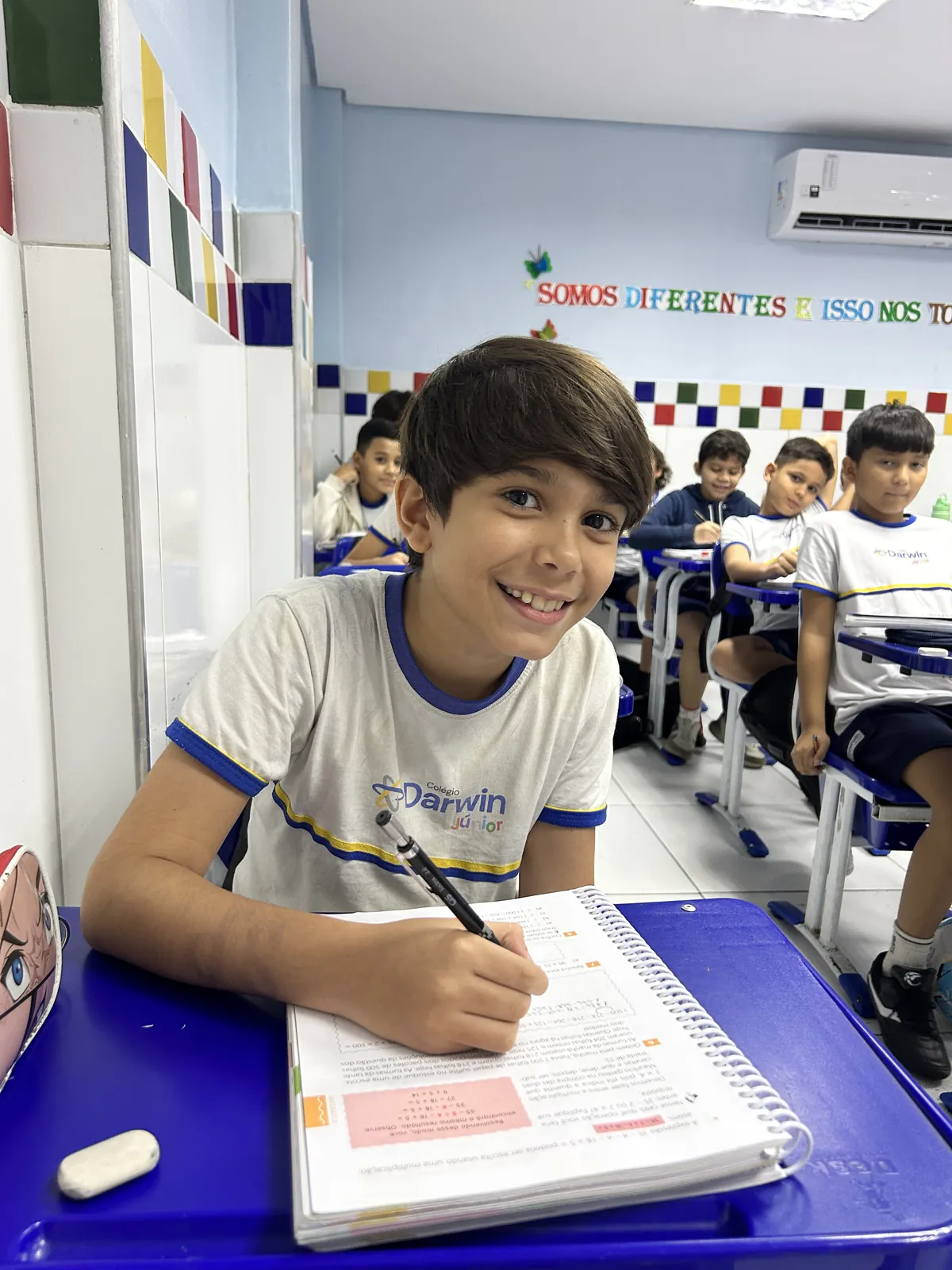 Aluno sorrindo em sala com o mural 'Somos diferentes e isso nos torna'.
