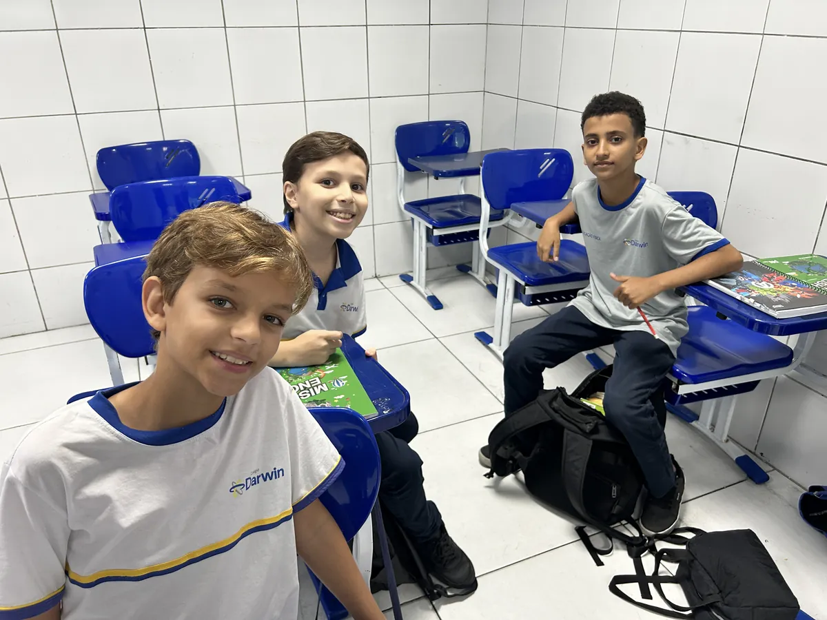 Três alunos sorrindo em sala com carteiras azuis do Darwin.