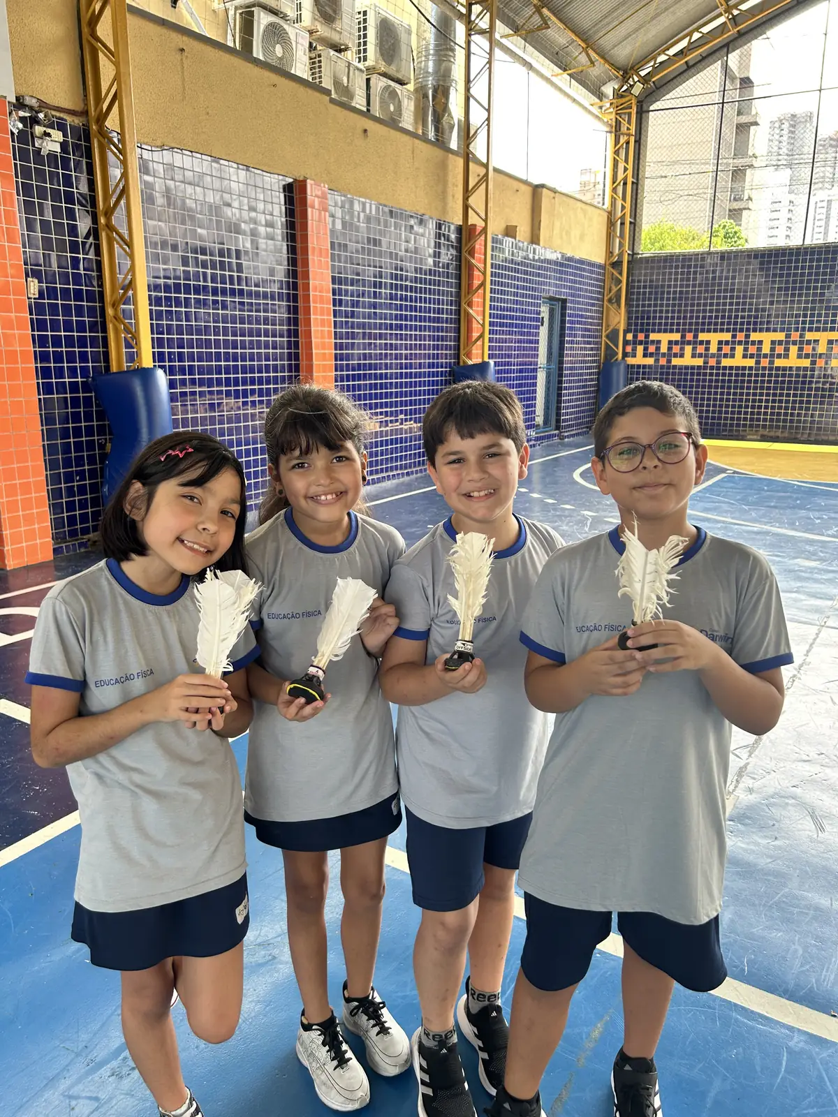Quatro alunos com penas brancas em atividade lúdica na quadra.