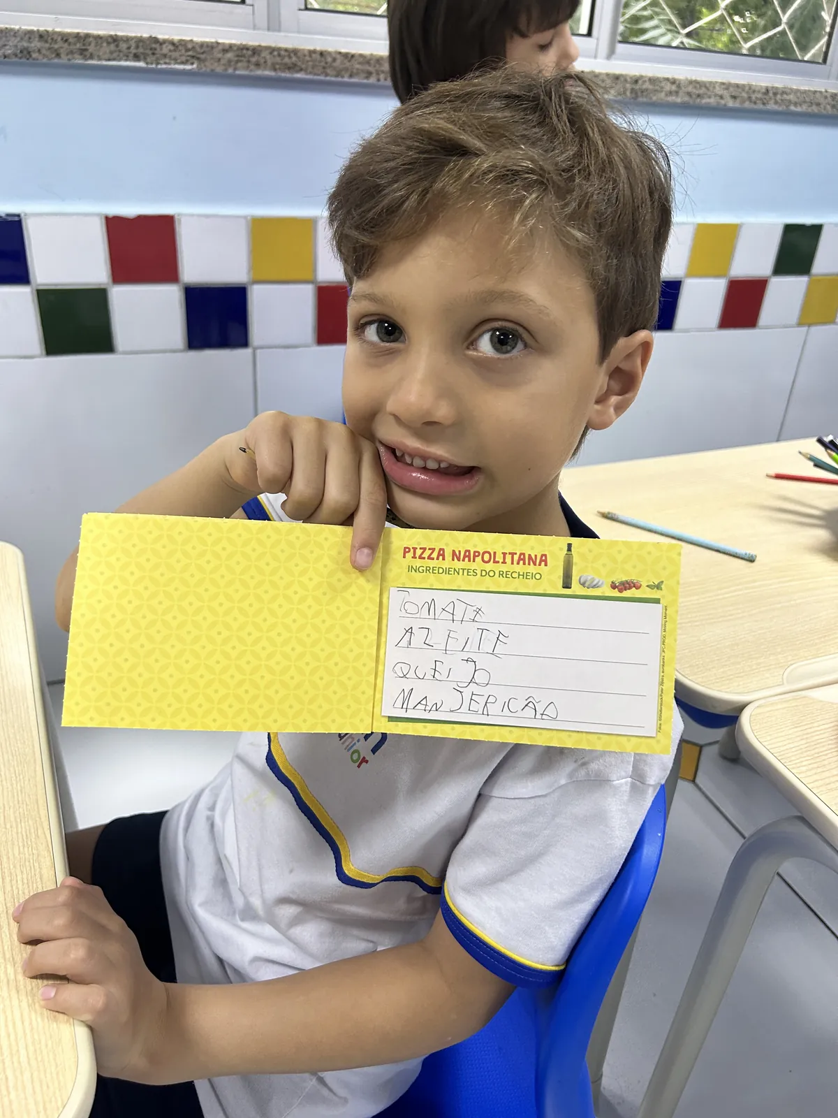 Menino do Infantil mostrando placa amarela com o nome de uma receita.