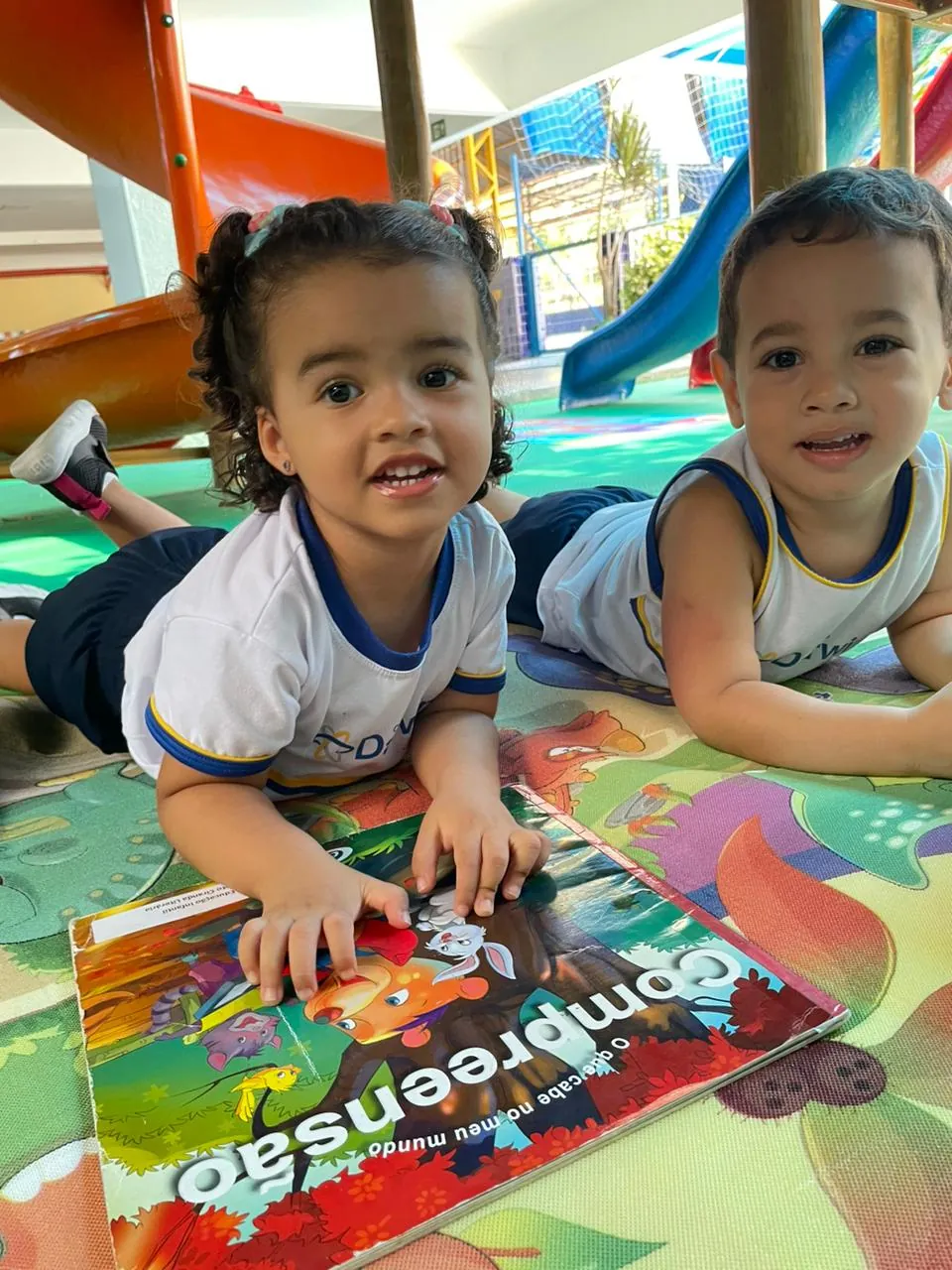 Dois alunos do Infantil deitados no tapete colorido explorando livro.