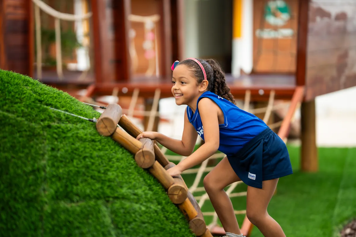 Aluna do STI escalando estrutura de madeira do playground com apoio das mãos e dos pés.