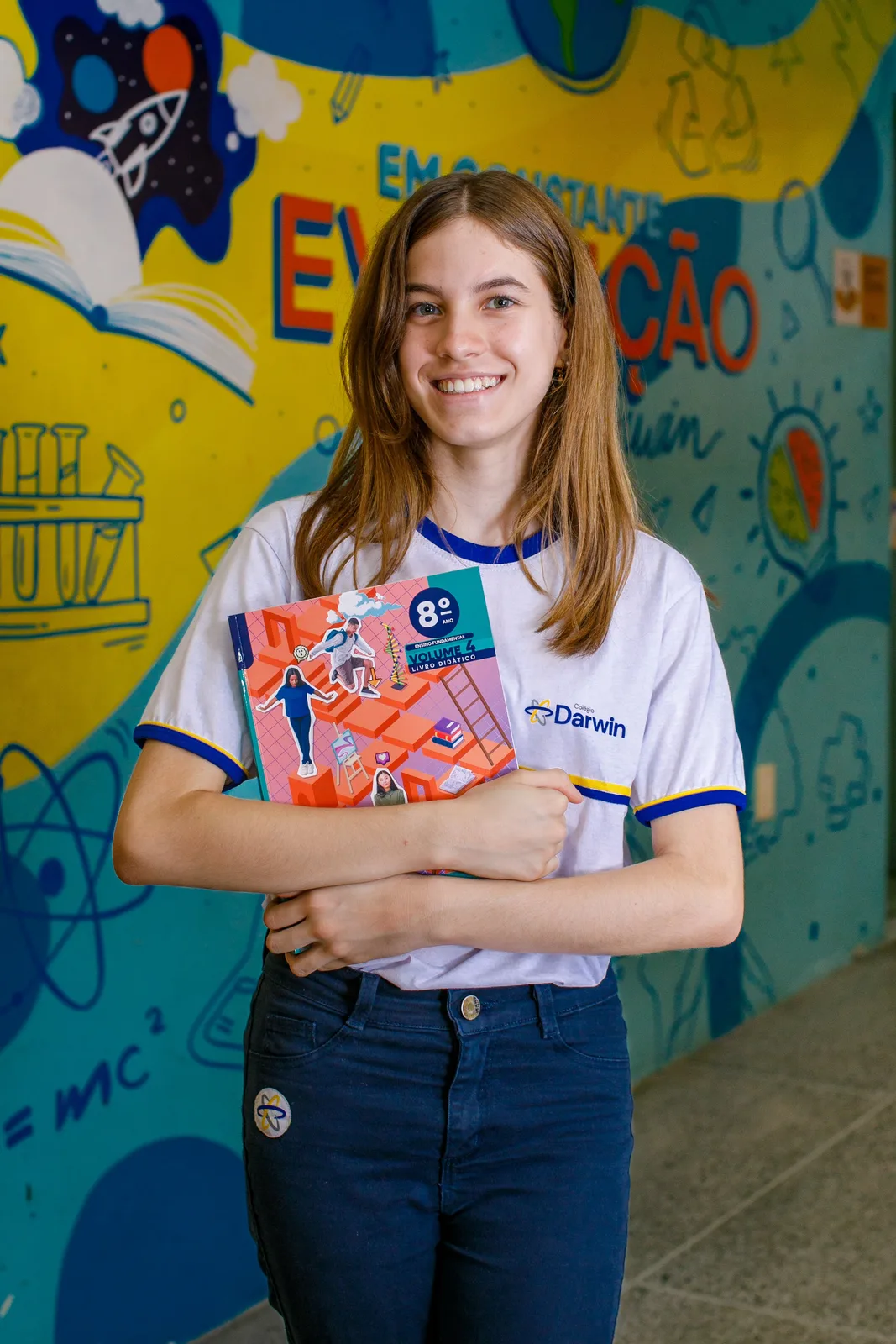 Aluna dos Anos Finais da Sede Aldeota abraçada aos livros do 8º ano, diante do mural grafitado do pátio
