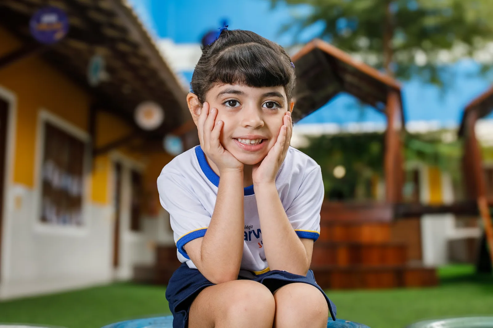 Criança da Educação Infantil do Darwin sentada no gramado do parquinho, com as mãos no rosto, sorrindo para a câmera.